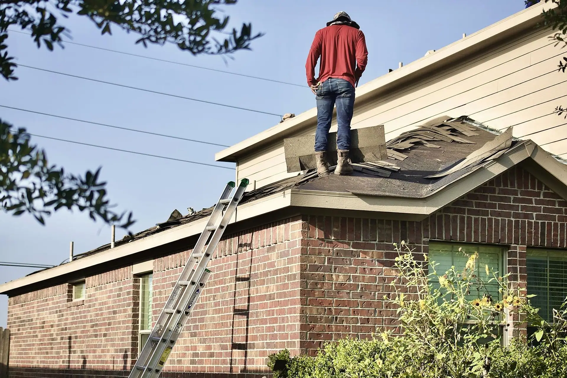 Roofing contractor standing on the ridge of a suburban home with a ladder set against the eaves