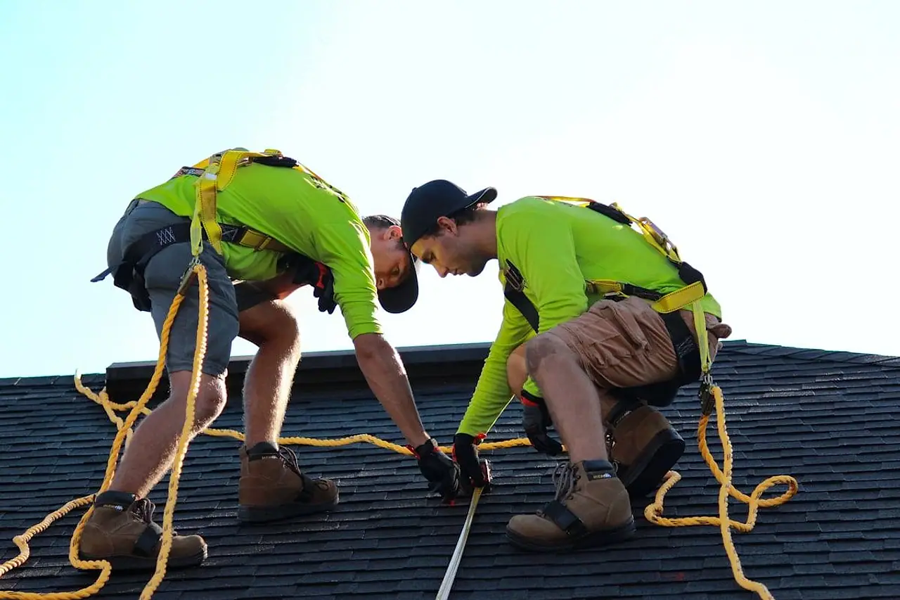 Two roofers fastening shingles on a residential roof, both wearing fall-protection harnesses